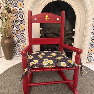 May include: A small red wooden rocking chair featuring a painted yellow cartoon bird and white flowers on the top back slat. The chair includes a removable black fabric seat cushion decorated with a white daisy pattern and tied at the corners.