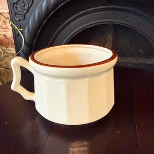 May include: Cream-colored ceramic mug with a brown rim and handle. The mug has a geometric, faceted design on its exterior. The handle is square and angular. The mug is set against a dark background.