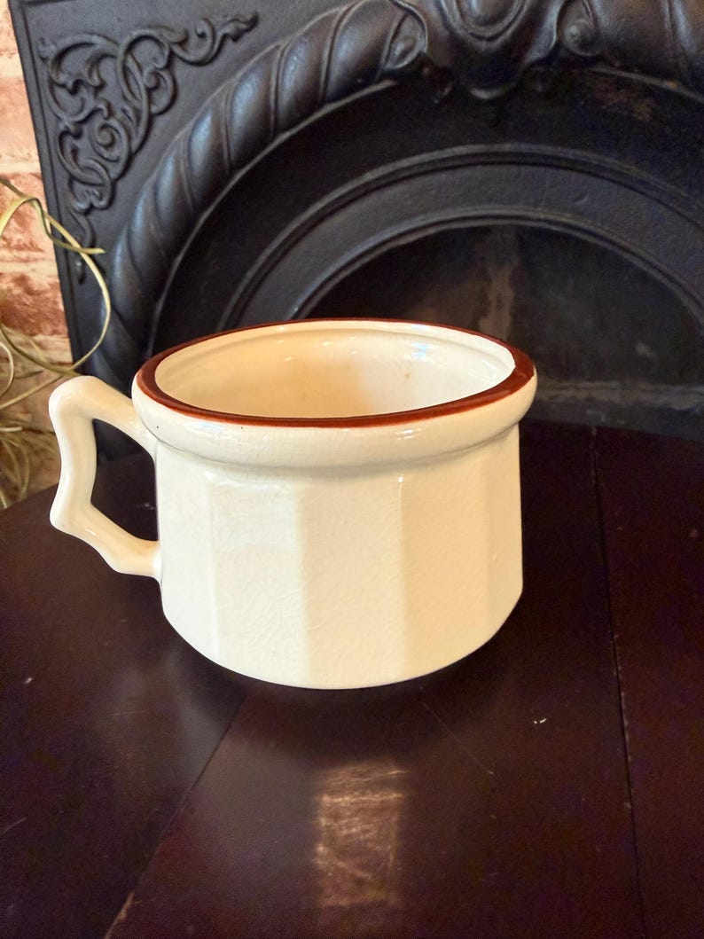 May include: Cream-colored ceramic mug with a brown rim and handle. The mug has a geometric, faceted design on its exterior. The handle is square and angular. The mug is set against a dark background.