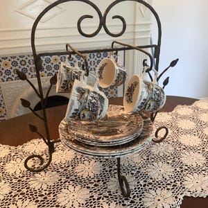 May include: A decorative wrought iron tea cup and saucer stand holding four cups and saucers. The cups and saucers have a black and white pastoral scene design. The stand is on a table with a cream-colored lace runner.
