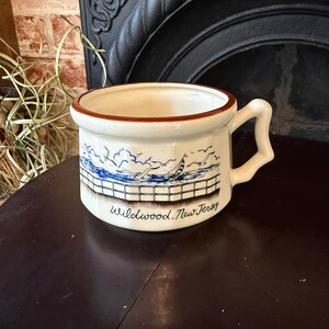 May include: Cream-colored ceramic mug with a brown rim and handle. The mug features a blue and white seaside scene with a sailboat and the text "Wildwood, New Jersey". The mug is sitting on a dark wooden surface.