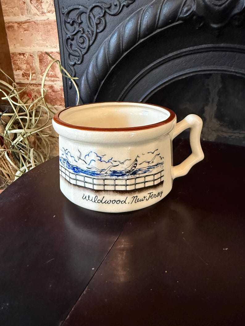 May include: Cream-colored ceramic mug with a brown rim and handle. The mug features a blue and white seaside scene with a sailboat and the text "Wildwood, New Jersey". The mug is sitting on a dark wooden surface.