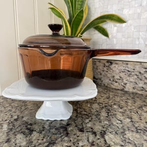 May include: A vintage amber-colored glass saucepan with a lid and handle, resting on a white, ornate cake stand. The saucepan is displayed in front of a white wall and a granite countertop, with a potted plant in the background.