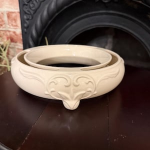 May include: Cream-colored, two-piece decorative bowl with ornate floral detailing. The outer bowl has a rounded shape with a decorative base, and the inner bowl is a smaller, circular insert. The bowls are made of ceramic and sit on a dark wooden surface.