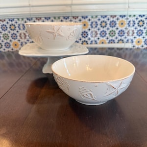 May include: Two off-white ceramic bowls with embossed seashell and starfish designs. One bowl sits on a white pedestal, the other on a dark wooden surface. The bowls have a rustic, coastal theme.
