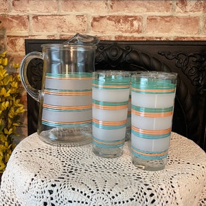 May include: A vintage glass pitcher and two matching glasses. The pitcher has a handle and a spout. The glasses and pitcher are decorated with horizontal stripes in frosted white, teal, and orange. The set is displayed on a white crocheted tablecloth.