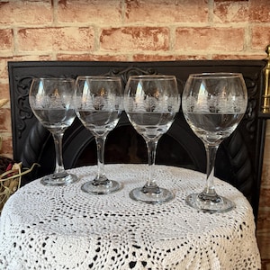 May include: Four clear wine glasses with a tree design etched on the bowl. The glasses are on a white, crocheted tablecloth. The background includes a brick wall and a black fireplace.