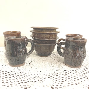 May include: A collection of handmade ceramic mugs and bowls. The mugs are dark brown with textured surfaces and handles. The bowls are stacked in the center, also in shades of brown. The items are arranged on a white, lace-like surface.