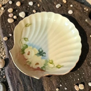 May include: A seashell-shaped dish with a scalloped edge and a painted floral design. The dish is primarily cream-colored with a gold trim. The floral design features white roses with green leaves and blue accents.