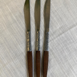 May include: Three stainless steel dinner knives with brown wooden handles. Each knife features a silver-toned metal bolster decorated with an engraved scrollwork pattern that transitions into a smooth, dark brown wooden handle grip.