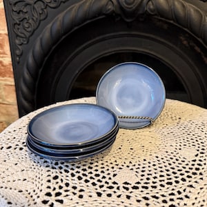 May include: A stack of four blue ceramic bowls with a dark blue rim and a single bowl on a metal stand. The bowls have a light blue interior and are set on a white crocheted tablecloth.