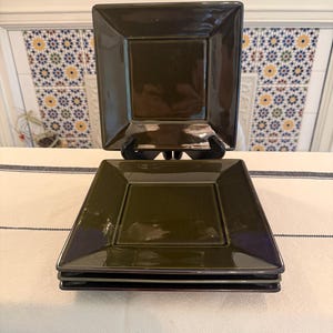 May include: A set of three square, dark olive green ceramic plates with a glossy finish. The plates feature a flat center and a wide, angled rim. One plate is displayed upright on a black stand, while two others are stacked horizontally below it.