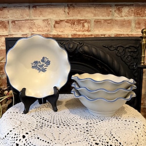 May include: Cream-colored ceramic bowls and a plate with a blue floral design and scalloped edges. The plate is on a black stand. Three bowls are stacked together. The items are on a white crocheted tablecloth.