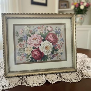 May include: Framed floral print featuring a bouquet of pink, red, and white peonies and other flowers. The artwork is set in a gold-colored frame with a rope-like border. The framed print is displayed on a dark wooden table with a lace doily.