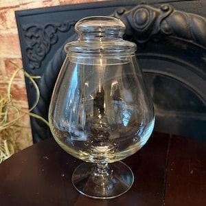 May include: A clear glass apothecary jar with a rounded body, a stemmed base, and a matching lid. The jar is empty and sits on a dark wooden surface, with a brick wall and a decorative black metal element in the background.
