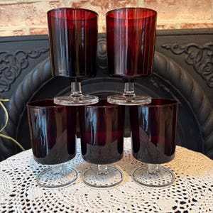 May include: A set of six vintage wine glasses. The glasses are a deep burgundy color with a clear stem and base. The glasses are arranged in a pyramid shape on a white lace doily.