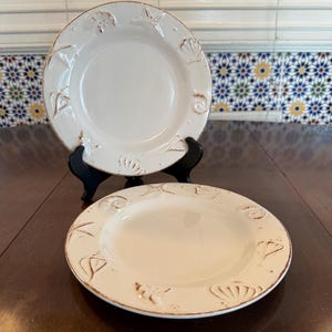 May include: Two off-white ceramic plates with a brown rim and embossed seashell designs. The plates are on a dark brown wooden table, with one plate on a black stand. The plates have a rustic, coastal theme.