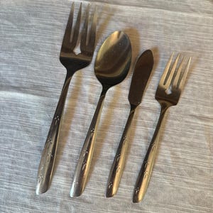 May include: A set of four silver-toned stainless steel utensils including a large three-tined fork, a soup spoon, a butter knife, and a smaller three-tined fork. Each handle features a repeating etched leaf pattern along the center stem.