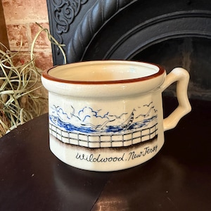 May include: Cream-colored ceramic mug with a brown rim and handle. The mug features a blue and white illustration of a seascape with a sailboat, seagulls, and a fence. The text "Wildwood, New Jersey" is printed below the illustration.