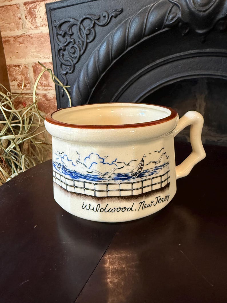 May include: Cream-colored ceramic mug with a brown rim and handle. The mug features a blue and white illustration of a seascape with a sailboat, seagulls, and a fence. The text "Wildwood, New Jersey" is printed below the illustration.