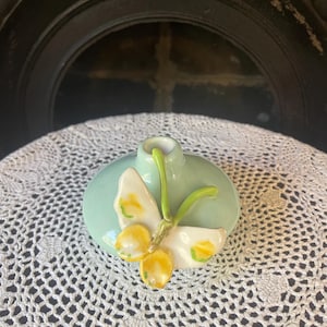 May include: A small, round, light-green ceramic vase with a white and yellow floral design. The vase has a single opening and is placed on a white, crocheted doily. The background is a dark, ornate structure.