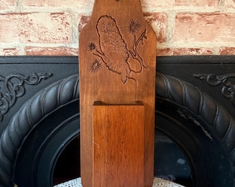 May include: A wooden match holder with a carved owl design. The match holder is rectangular with a small pocket at the bottom. The owl is perched on a branch with detailed feathering. The wood has a warm, reddish-brown tone.