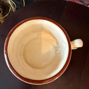 May include: A ceramic mug with a cream-colored interior and a brown rim and handle. The mug is set on a dark brown surface. The mug is empty.