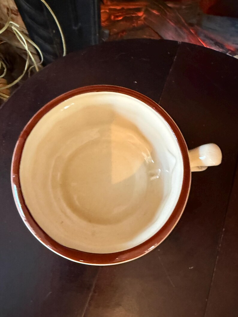 May include: A ceramic mug with a cream-colored interior and a brown rim and handle. The mug is set on a dark brown surface. The mug is empty.