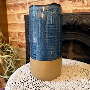 May include: A cylindrical ceramic vase with a two-tone design. The upper portion is a glossy, textured blue, while the lower section is a matte tan. The vase has a slightly irregular rim and is likely handmade. It is sitting on a white lace doily.