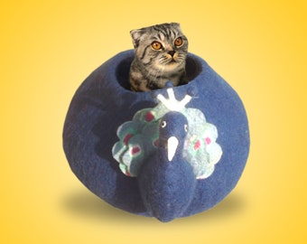 May include: A dark blue felted wool cat cave shaped like a peacock with a rounded body, a protruding neck, and a fan-shaped tail featuring light blue, white, and pink accents. A grey tabby cat with orange eyes is sitting inside the circular opening.