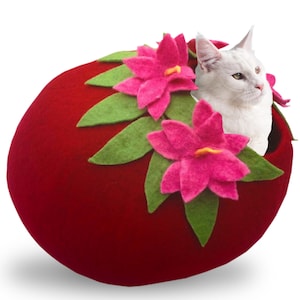 May include: A red felt cat bed shaped like a sphere with pink flowers and green leaves around the opening. A white cat is sitting inside the bed.