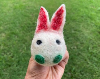 May include: A white felt bunny with red ears and green felt eyes. The bunny is held in a hand.
