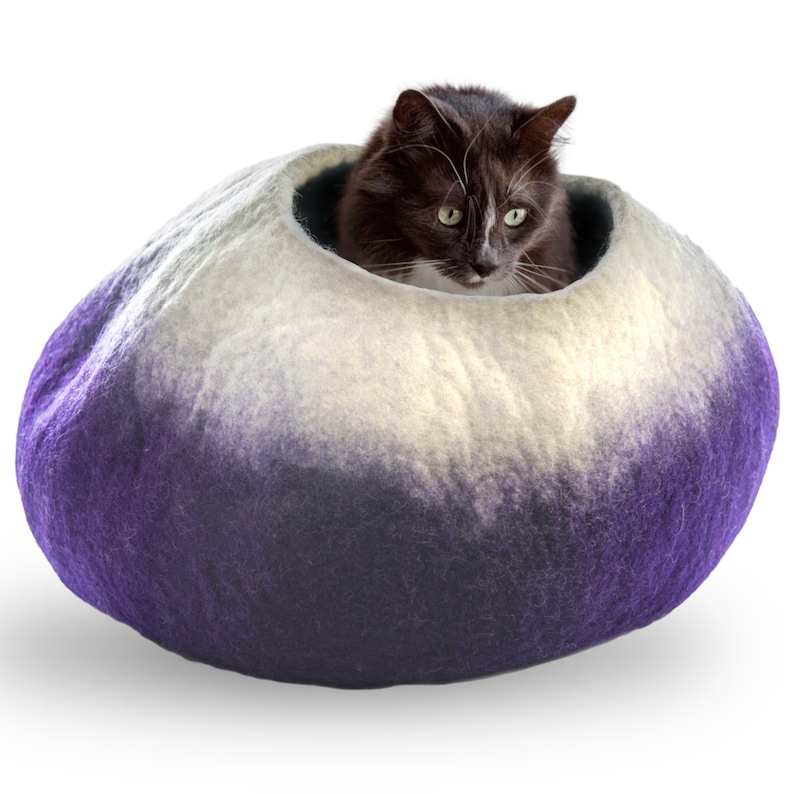 May include: A purple and white felted cat bed with a cat inside. The bed is shaped like a large, rounded stone.