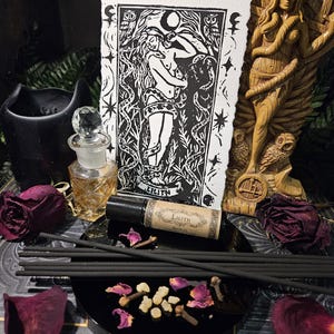 May include: A black and white print of Lilith, a perfume bottle, incense sticks, and dried rose petals are arranged on a black tray. A wooden statue and a black candle holder are in the background. The perfume bottle has a clear glass stopper.