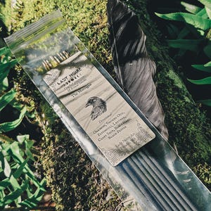 May include: A clear plastic bag containing black incense sticks with a tan label featuring the text Lady Noire, The Morrigan, and a raven illustration. The label lists ingredients including oakmoss, smoked oud, patchouli, leather, clary sage, blackberry, and black pepper. A dark feather is placed next to the bag.