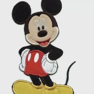 May include: Embroidered Mickey Mouse patch featuring a smiling face with black ears, a tan face, and a red mouth. Mickey is wearing white gloves with red shorts and yellow shoes. The patch is designed for application on clothing or accessories.