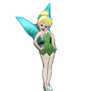 May include: Embroidered patch featuring a cartoon fairy with blonde hair, green dress, and teal wings. The fairy is in a standing pose with her hands on her hips. The patch is designed with detailed stitching.