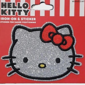 May include: A glittery silver Hello Kitty iron-on and sticker patch with a red bow. The patch has a black outline and is on a red background with white stripes. The text "HELLO KITTY" is at the top.