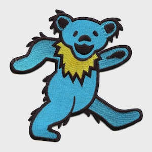 May include: Embroidered patch featuring a stylized blue bear with a yellow collar and black outlines. The bear is depicted in a dancing pose, with its arms and legs outstretched. The patch is likely designed to be ironed on or sewn onto clothing or accessories.