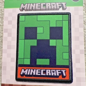 May include: A Minecraft iron-on patch featuring a pixelated Creeper face in green and dark blue. The patch is rectangular with a black border and the Minecraft logo in white on an orange background. The patch is on a green and brown backing.