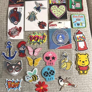 May include: A collection of assorted embroidered iron-on patches featuring various designs including Spider-Man, insects, a heart, a Jaws Amity Island logo, a gumball machine, a blue anchor, a Smurf, butterflies, skulls, a banana, Winnie the Pooh, and text.