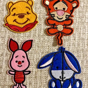 May include: Four embroidered patches featuring characters from Winnie the Pooh. Includes Winnie the Pooh in yellow, Tigger in orange, Piglet in pink, and Eeyore in blue. Each patch has a black outline and is on a beige background.