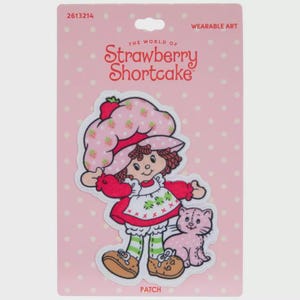 May include: A Strawberry Shortcake patch on a pink backing. The patch features a cartoon illustration of Strawberry Shortcake in a red dress and hat, with a pink cat. The text "The World of Strawberry Shortcake" is at the top.