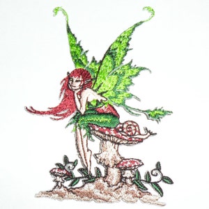 May include: Embroidered patch featuring a fairy with red hair, green wings, and a red and green dress. The fairy is seated on a mushroom with a snail. Additional mushrooms and foliage are at the base.
