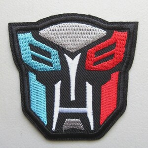 May include: Embroidered patch featuring a stylized emblem in black, with blue, red, white, and silver accents. The patch has a black border and a geometric design, suitable for application on clothing or accessories.