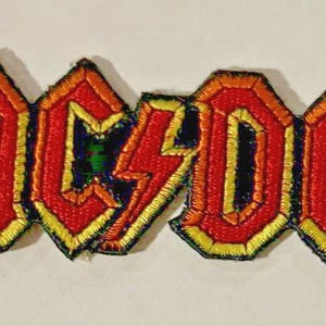 May include: Embroidered patch featuring the AC/DC logo in red, orange, and yellow. The design includes the band's name in a stylized font with a lightning bolt in the center. The patch is likely for application on clothing or accessories.