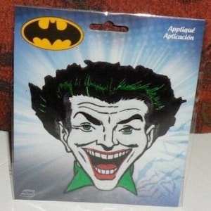 May include: A fabric patch featuring the face of the Joker with black hair, green highlights, white skin, and a wide red-lipped grin. The patch is displayed on a blue and white backing card with the yellow and black Batman logo at the top left.