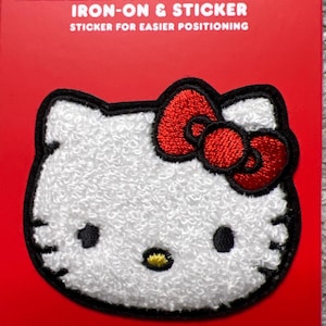 May include: A Hello Kitty iron-on and sticker patch. The patch features a white, textured Hello Kitty face with black eyes, a yellow nose, and a red bow. The patch is outlined in black and set against a red background.