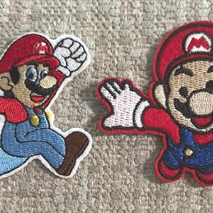 May include: Two embroidered fabric patches featuring the character Mario. The left patch shows Mario running with a raised fist, wearing a red hat with an M, red shirt, blue overalls, and brown shoes. The right patch shows Mario jumping with arms outstretched.