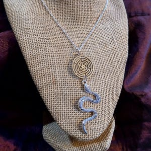 Op de afbeelding: Een zilverkleurige ketting met een hanger. De hanger heeft een goudkleurig cirkelvormig labyrintontwerp en een gedetailleerde slangenbedel. De ketting wordt gepresenteerd op een met jute bedekte buste tegen een donkere achtergrond.
